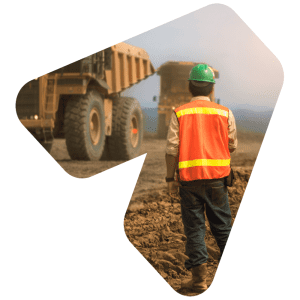 A miner wearing a high visibility vest and hardhat watches two large mining trucks drive in the background